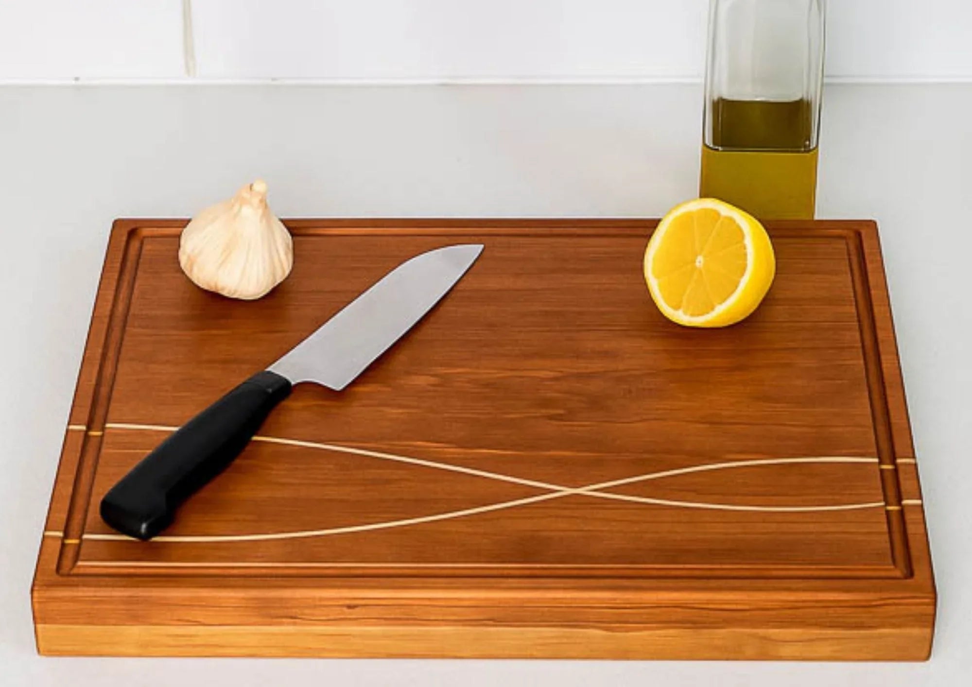 Edge grain wooden cutting board with knife, garlic bulb, lemon half, and olive oil bottle