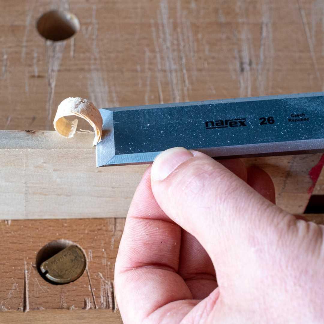 Hand holding Narex 26 chisel carving a thin wood shaving on a workbench