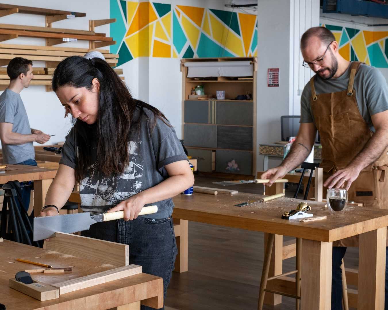 Two people woodworking with hand tools in a bright workshop with geometric wall art
