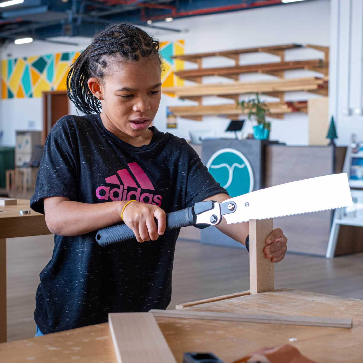 Young girl sawing wood in a brightly lit workshop with woodworking tools and shelves in background
