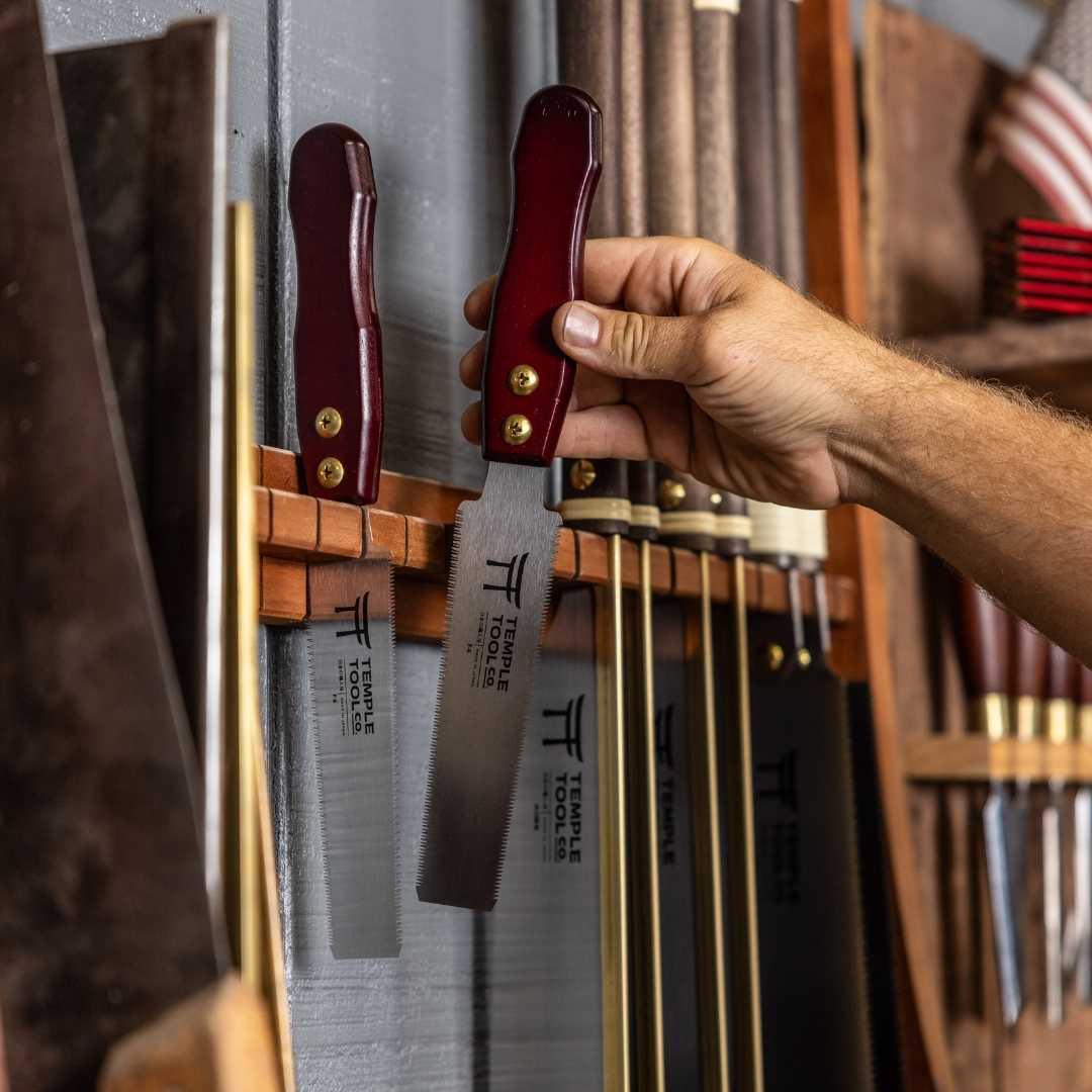 Hand holding a Temple Tool flush cut pull saw with red handle on wooden rack