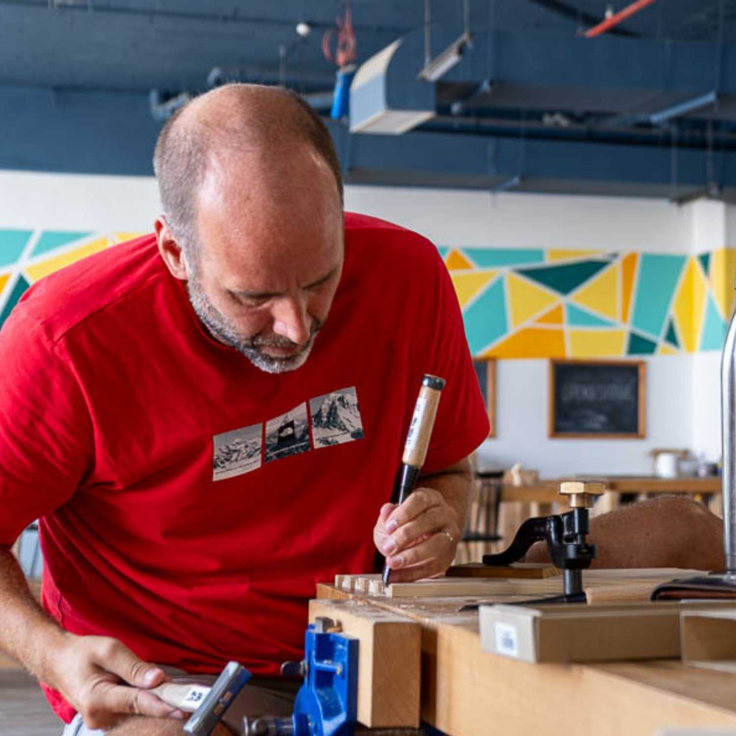 A man in a red t-shirt focused on a woodworking task, using a chisel and a wooden mallet to carve joinery into a piece of timber. The wood is secured in a blue metal vice on a heavy timber workbench within a bright workshop featuring geometric wall art.