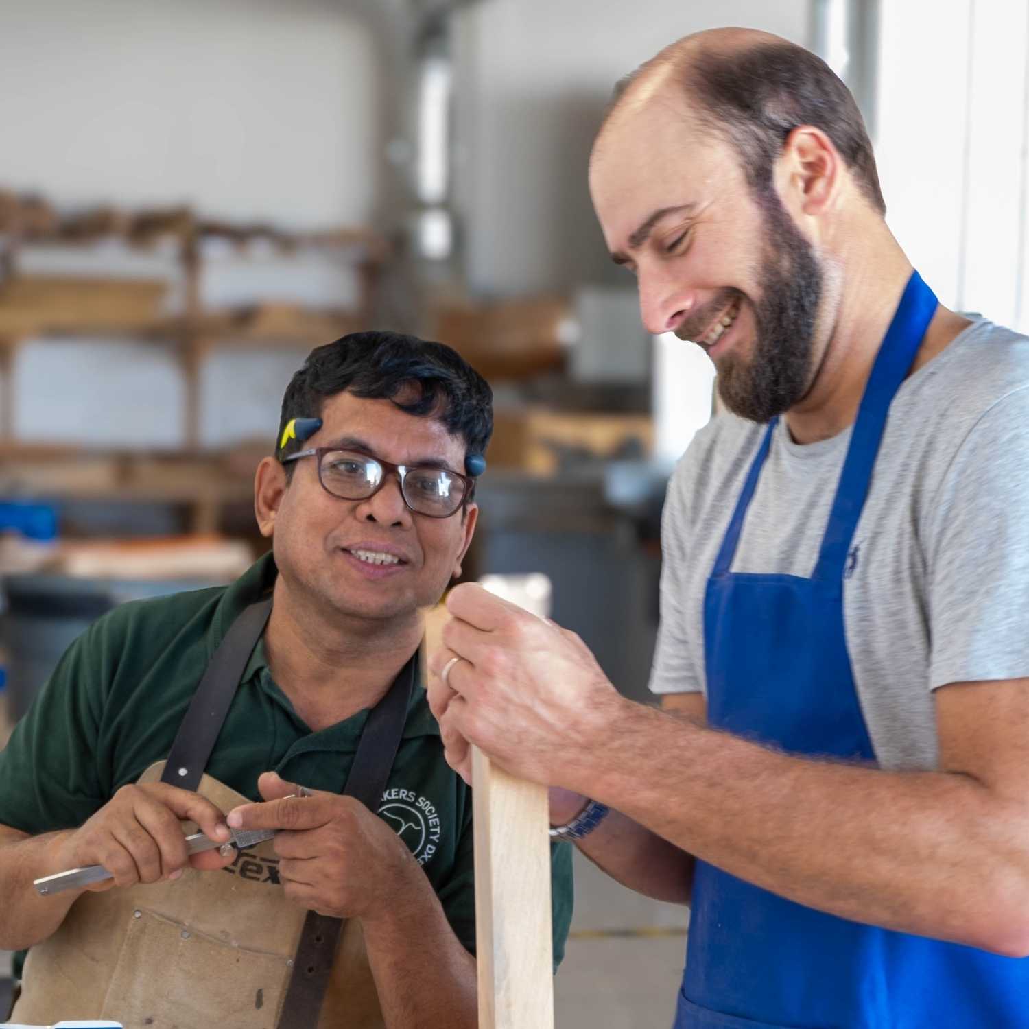 A woodworking student in a blue apron and a bearded instructor in a tan "Narex" apron share a smile while inspecting a piece of wood. The instructor holds a measuring tool as they discuss the progress of a joinery project in a professional workshop setting.